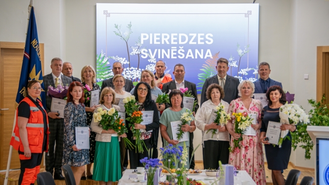 NMPD darbinieki, kuri nostrādājuši vairāk nekā 25 gadus stāv uz skatuves, svinīgā pasākumā, fonā ekrānā rakstīts "Pieredzes svinēšana"