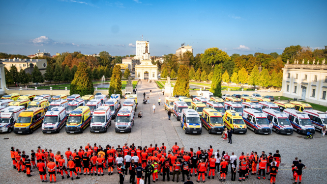 Skats no augšas, kur redzamas daudz dažādu ārvalstu ambulances un priekšā stāv daudz mediķu un paramediķu oranžās formās