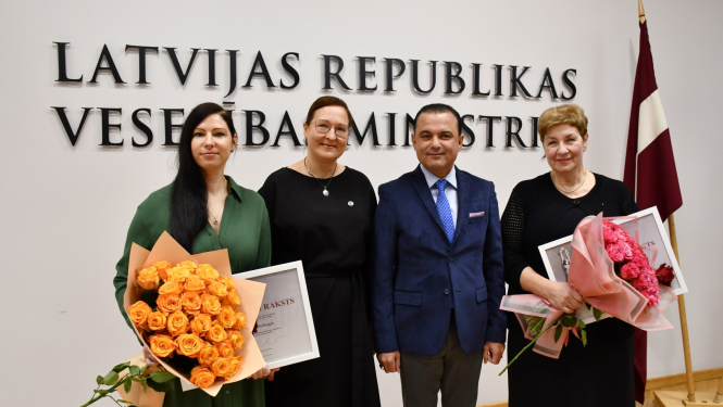 Cilvēki stāv un fonā uz sienas ministrijas nosaukuma uzraksts un Latvijas karogs