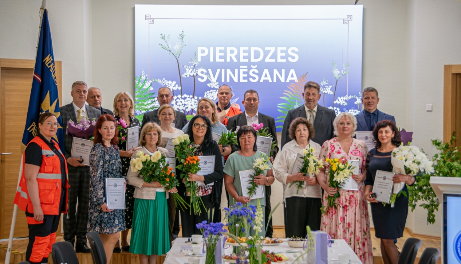 NMPD darbinieki, kuri nostrādājuši vairāk nekā 25 gadus stāv uz skatuves, svinīgā pasākumā, fonā ekrānā rakstīts "Pieredzes svinēšana"