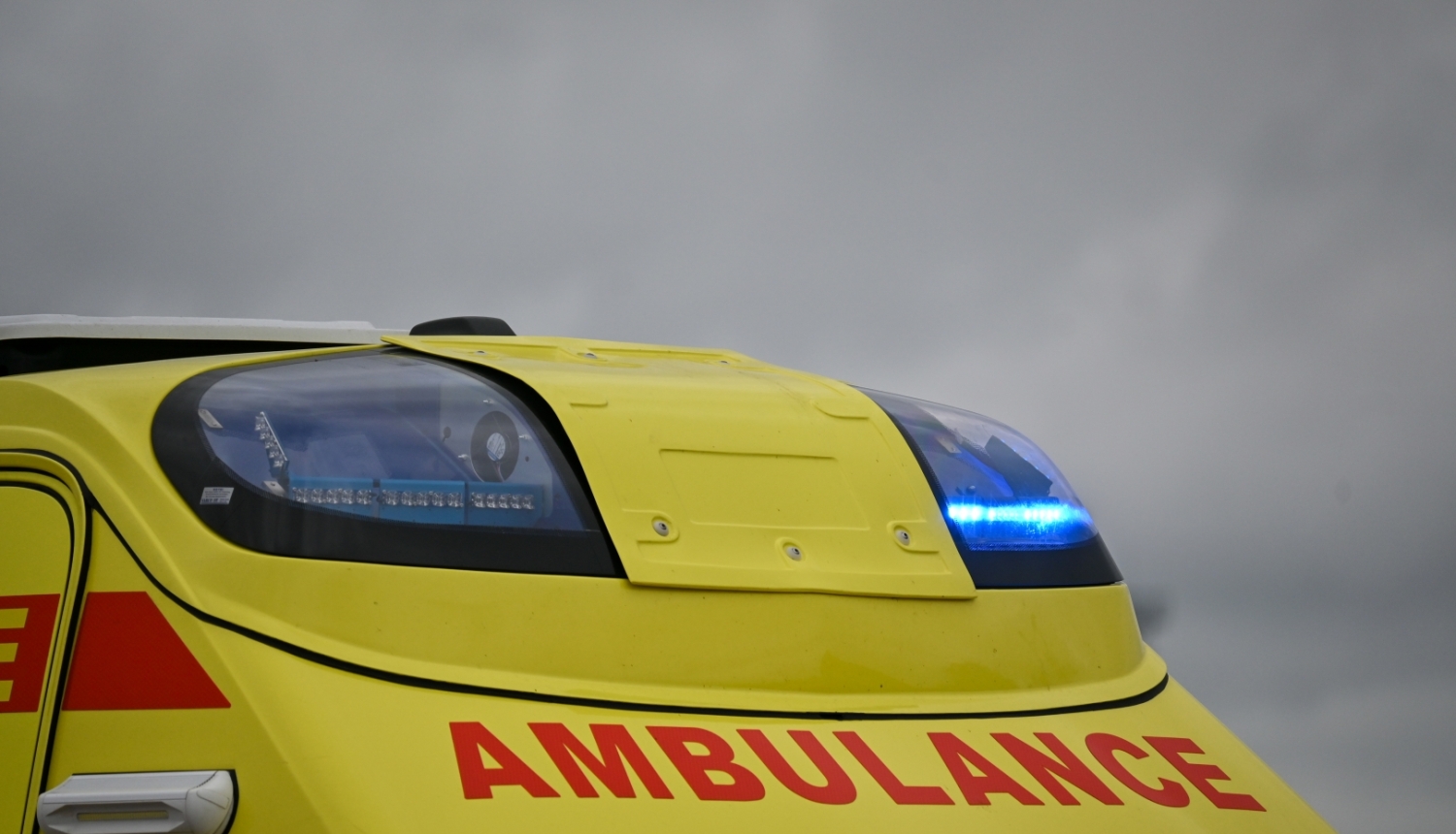 NMPD operatīvais auto, tuvplānā bākugunis un uzraksts AMBULANCE. Fonā tumšas debesis.