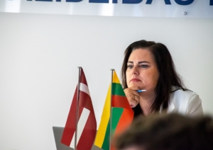 Sieviete baltā žaketē un uz galda stāv LT un LV karogi