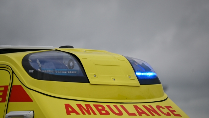 NMPD operatīvais auto, tuvplānā bākugunis un uzraksts AMBULANCE. Fonā tumšas debesis.
