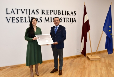 Dita Heiberga saņem atzinības rakstu no veselības ministra. Fonā uz sienas ministrijas nosaukuma uzraksts un Latvijas, Eiropas karogi