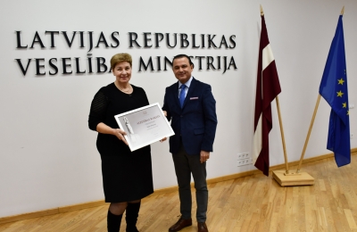 Maija Bušmane saņem atzinības rakstu no veselības ministra. Fonā uz sienas ministrijas nosaukuma uzraksts un Latvijas, Eiropas karogi