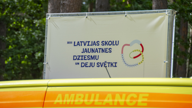 Baneris ar uzrakstu 13. Latvijas skolu jaunatnes dziesmu un deju svētki, priekšplānā ambulnaces mašīna ar uzrakstu