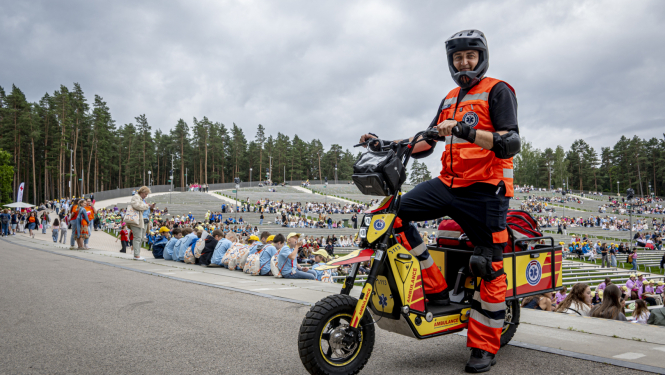 NMPD darbinieks formā un melnā ķiverē stāv pie NMPD elektroskrejriteņa un fonā Mežaparka estrāde