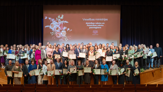 Veselības ministrijas apbalvojumu saņēmēji stāv uz skatuves un fonā ekrāns ar apbalvošanas pasākuma nosaukumu