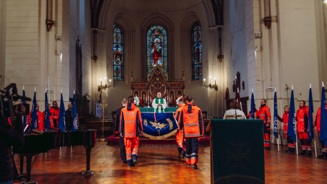 NMPD darbinieki nes dienesta svētku karogu baznīcā, apkārt mediķi formās ar struktūrvienību karogiem