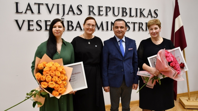 Cilvēki stāv un fonā uz sienas ministrijas nosaukuma uzraksts un Latvijas karogs