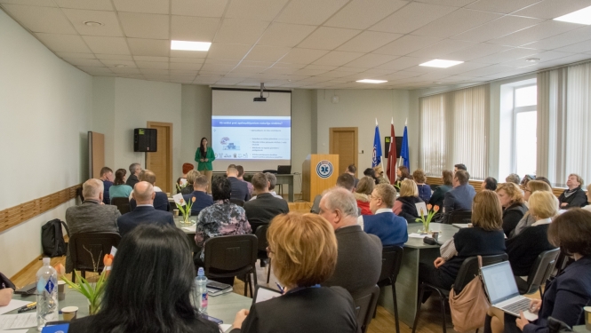 Cilvēki sēž pie galdiem un zāles galā redzama prezentācija