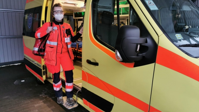 Mediķe NMPD formā un ar respiratoru, ar somu plecā stāv pie operatīvā auto