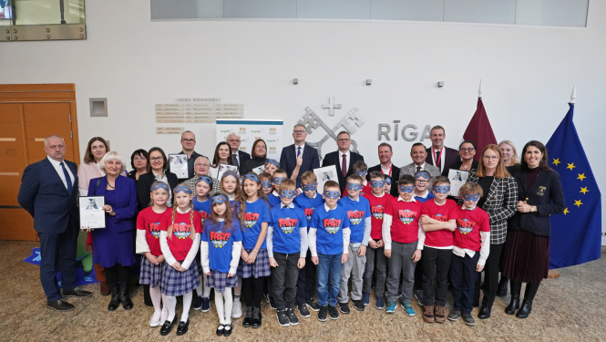 Bērni un dažādu iestāvju pārstāvji kopbildē, fonā Rīgas uzraksts