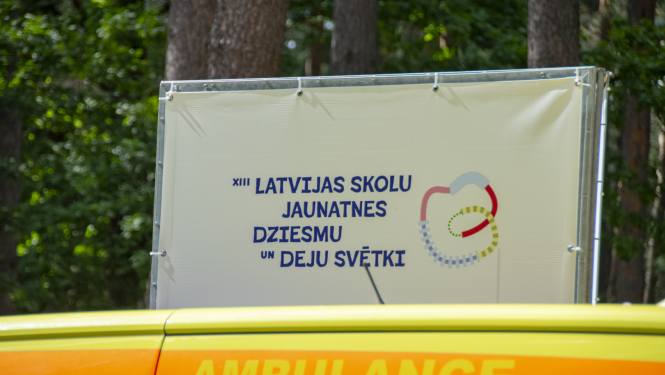 Baneris ar uzrakstu 13. Latvijas skolu jaunatnes dziesmu un deju svētki, priekšplānā ambulnaces mašīna ar uzrakstu