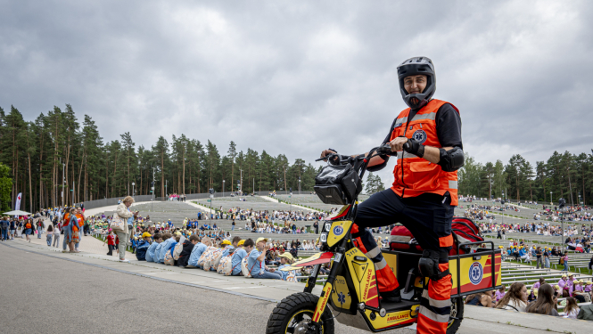 NMPD darbinieks formā un melnā ķiverē stāv pie NMPD elektroskrejriteņa un fonā Mežaparka estrāde