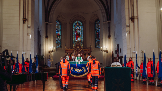 NMPD darbinieki nes dienesta svētku karogu baznīcā, apkārt mediķi formās ar struktūrvienību karogiem