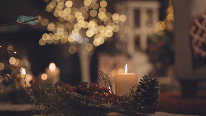 balta svece uz galda, uz galda čiekurs un zari, fonā redzamas dzeltenas gaismiņas