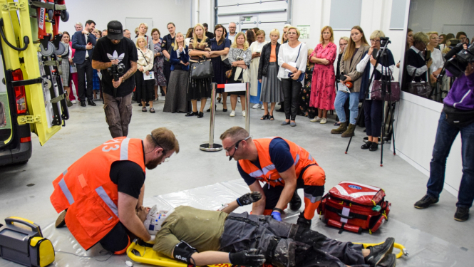 Mediķi mācību simulācijā sniedz palīdzību pacientam, fonā stāv cilvēki, kas vērā šo procesu