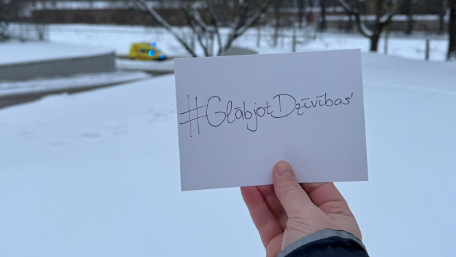 Rokās lapiņa ar uzrakstu #GlābjotDzīvības, apkārt sniegs un fonā brauc NMPD operatīvais auto