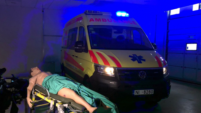 Manekens uz nestuvēm, fonā operatīvais auto ar ieslēgtām signālugunīm