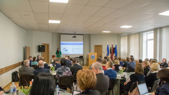 Cilvēki sēž pie galdiem un zāles galā redzama prezentācija