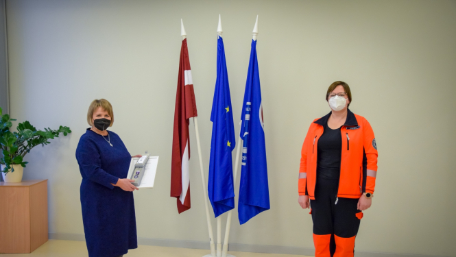 Stella Lapiņa ar NMPD direktori stāv pie valsts un dienesta karogiem