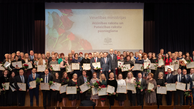 Cilvēki stāv uz skatuves, fonā ekrāns, kurā rakstīts - Veselības ministrijas Atzinības rakstu un Pateicības rakstu pasniegšana