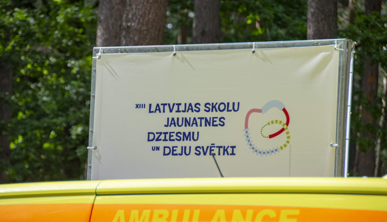 Baneris ar uzrakstu 13. Latvijas skolu jaunatnes dziesmu un deju svētki, priekšplānā ambulnaces mašīna ar uzrakstu