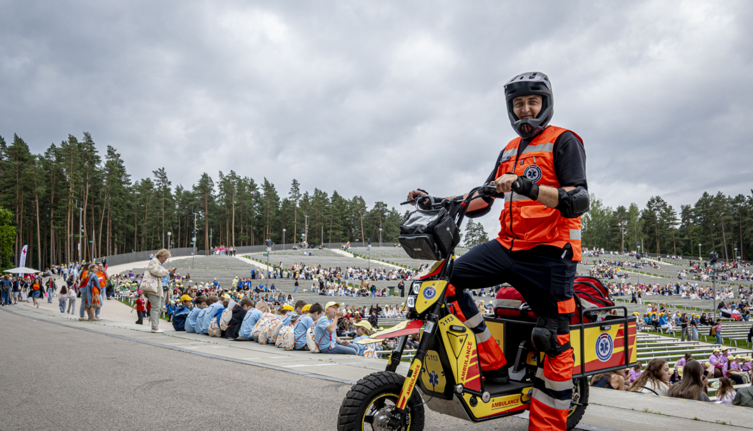 NMPD darbinieks formā un melnā ķiverē stāv pie NMPD elektroskrejriteņa un fonā Mežaparka estrāde