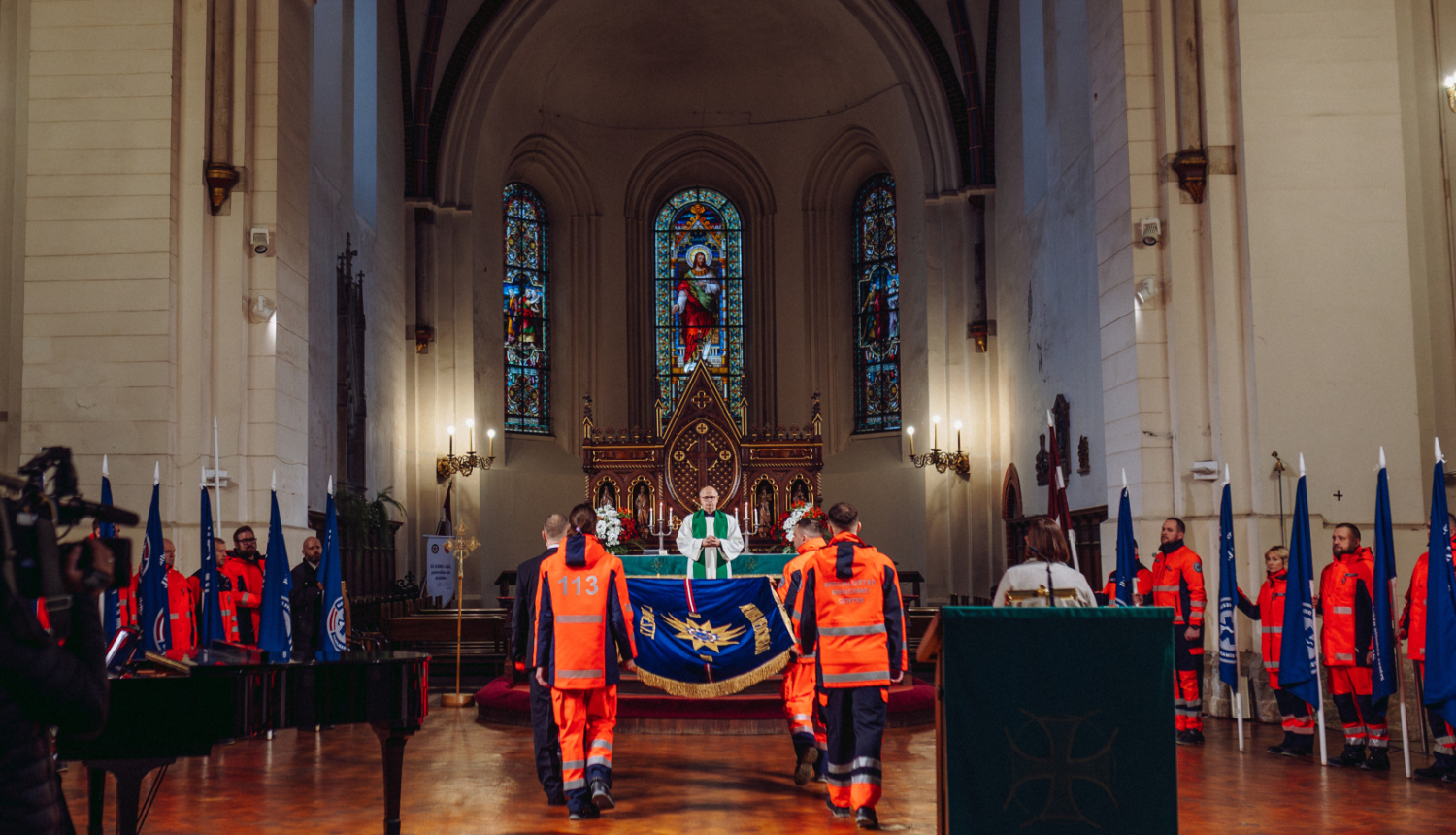 NMPD darbinieki nes dienesta svētku karogu baznīcā, apkārt mediķi formās ar struktūrvienību karogiem