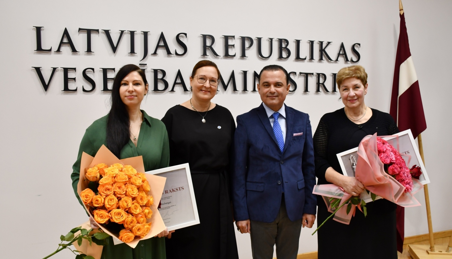 Cilvēki stāv un fonā uz sienas ministrijas nosaukuma uzraksts un Latvijas karogs