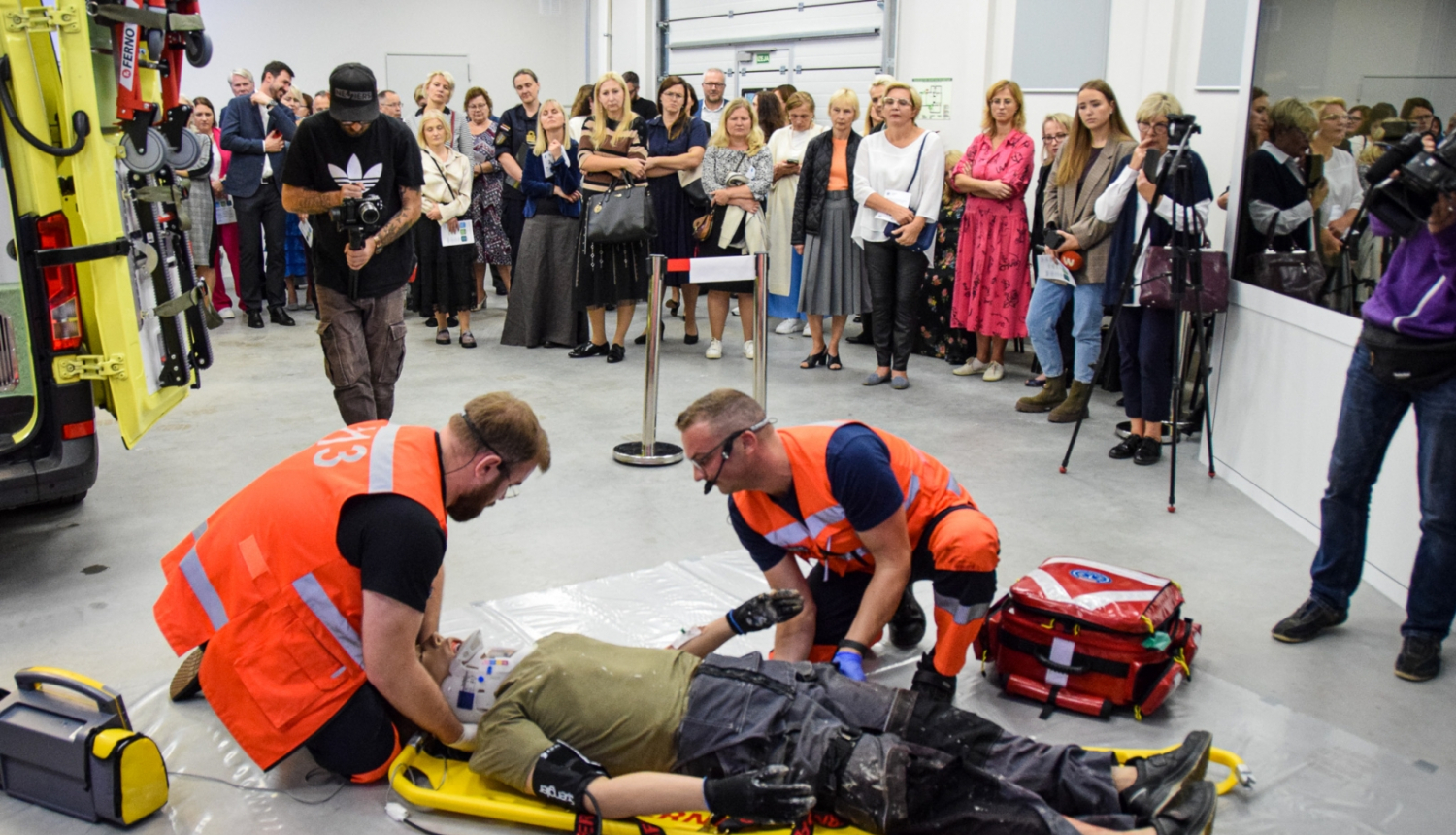 Mediķi mācību simulācijā sniedz palīdzību pacientam, fonā stāv cilvēki, kas vērā šo procesu