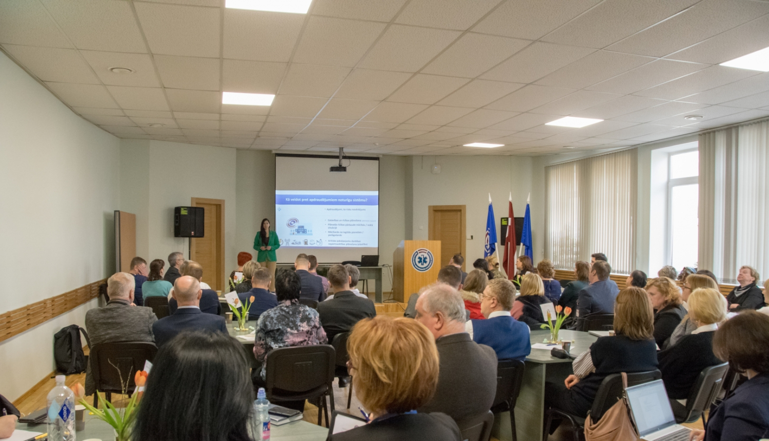 Cilvēki sēž pie galdiem un zāles galā redzama prezentācija