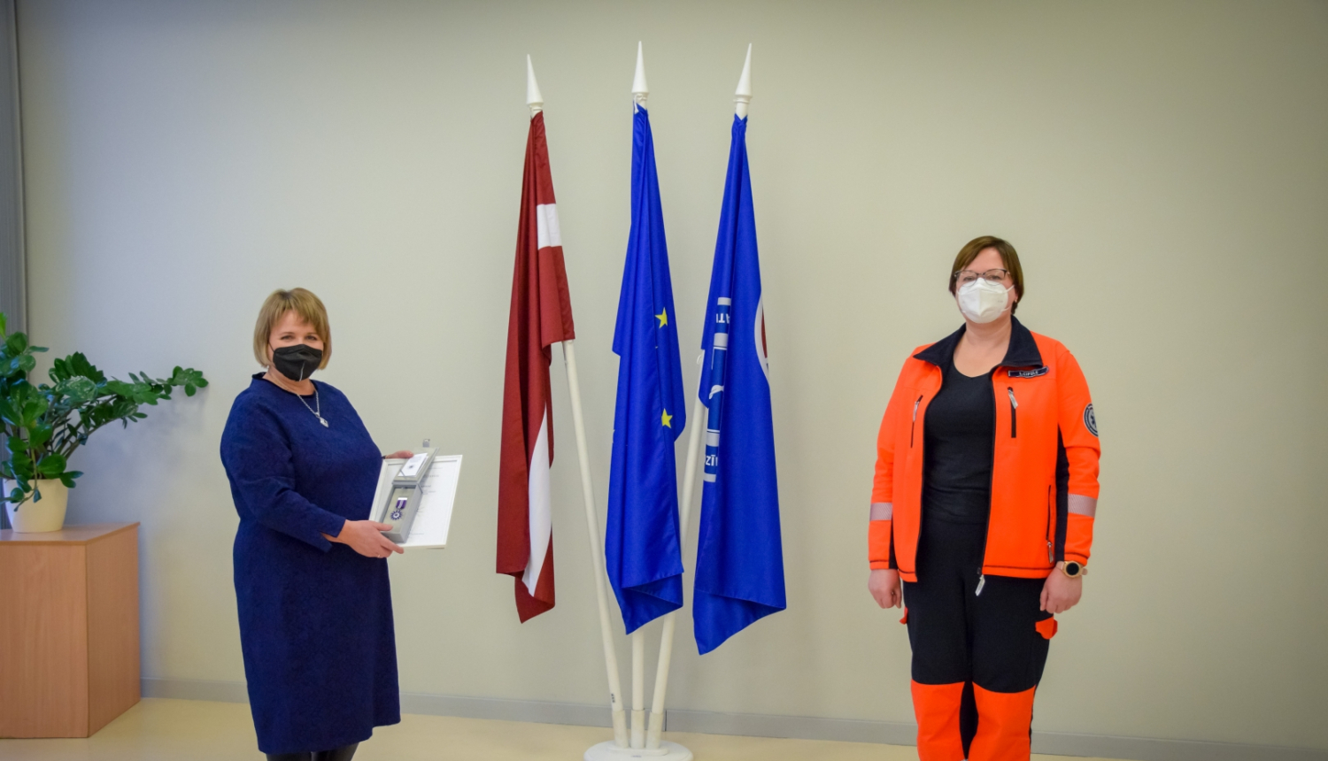 Stella Lapiņa ar NMPD direktori stāv pie valsts un dienesta karogiem