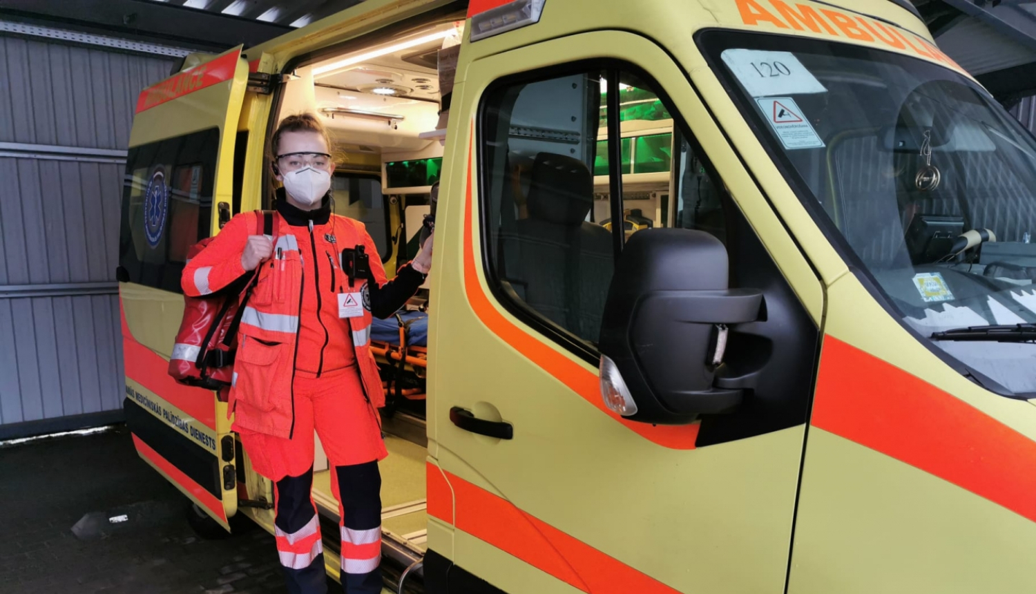 Mediķe NMPD formā un ar respiratoru, ar somu plecā stāv pie operatīvā auto