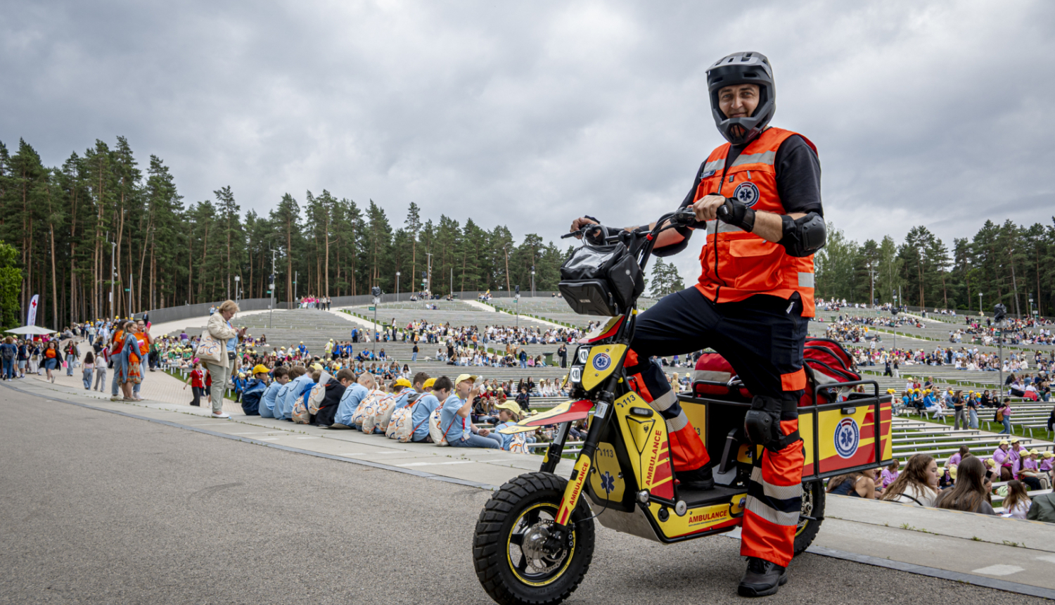 NMPD darbinieks formā un melnā ķiverē stāv pie NMPD elektroskrejriteņa un fonā Mežaparka estrāde