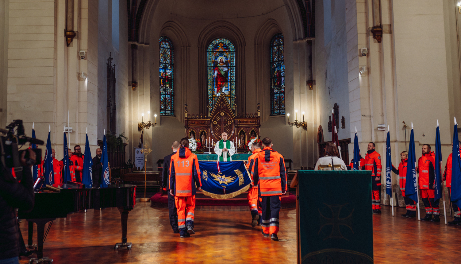 NMPD darbinieki nes dienesta svētku karogu baznīcā, apkārt mediķi formās ar struktūrvienību karogiem