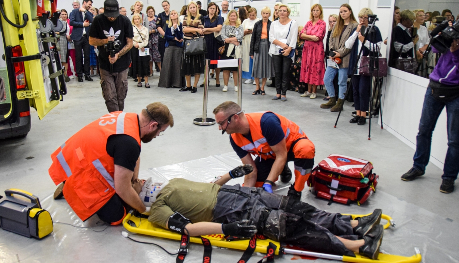 Mediķi mācību simulācijā sniedz palīdzību pacientam, fonā stāv cilvēki, kas vērā šo procesu