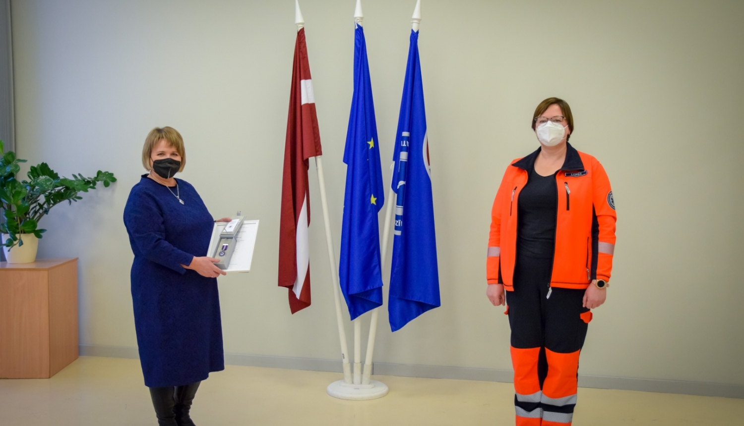 Stella Lapiņa ar NMPD direktori stāv pie valsts un dienesta karogiem