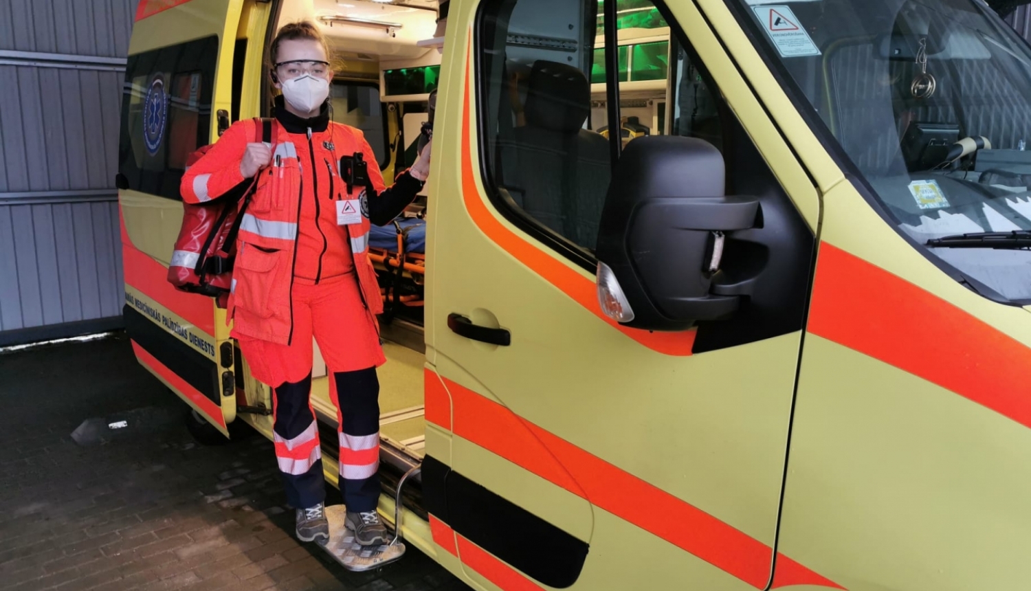 Mediķe NMPD formā un ar respiratoru, ar somu plecā stāv pie operatīvā auto