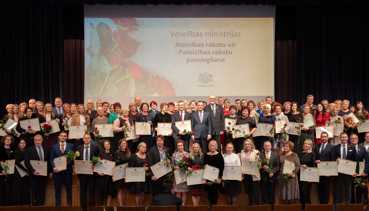 Cilvēki stāv uz skatuves, fonā ekrāns, kurā rakstīts - Veselības ministrijas Atzinības rakstu un Pateicības rakstu pasniegšana